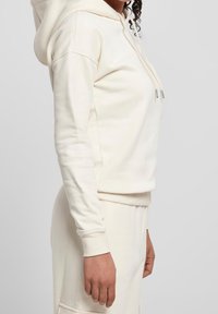 Sudadera con capucha de color crema hecha de tela suave, con un bolsillo frontal tipo canguro, puños acanalados y cierre con cordón en la capucha.