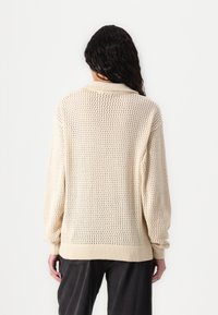 Strikket beige genser med åpen vevdesign, lange ermer, ribbestrikkede mansjetter og ribbestrikket nederkant. Har en klassisk krage.