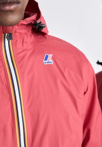 Rood lichtgewicht jack met een ritssluiting aan de voorkant, voorzien van een wit, zwart en geel streepdessin en een K-WAY-logo op de borst.