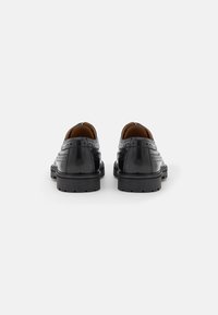 Zapatos de cuero negro con un acabado brillante, que presentan detalles de brogue perforados, suelas planas y talones redondeados. Diseño clásico con contraste de texturas.