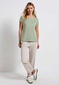 Camiseta verde claro de manga corta con un estampado plateado texturizado, combinada con pantalones de pierna ancha beiges y zapatillas blancas. Diseño sencillo y casual.