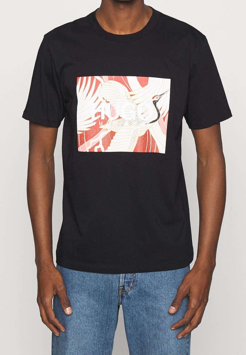 Sort T-shirt med et rød og hvidt kranegrafik og delvist synlig hvid tekst, båret af en person i blå jeans.