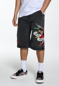 Shorts in denim nero con ricami che raffigurano un uccello, un cuore rosso e un banner con la scritta "VERA MORTE". Taglio dritto classico con tasche laterali.