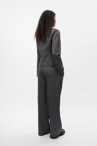 Žena s kučeravými vlasmi v sivom prúžkovanom obleku a čiernych topánkach, stojaca s rukami vreckách a chrbtom k divákovi na bielom pozadí.