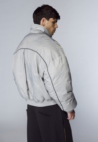 Uomo che indossa una giacca imbottita grigia chiara oversize con rifiniture nere, di spalle, abbinata a pantaloni neri su uno sfondo semplice.