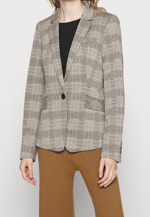 Personne portant un blazer à carreaux beige et marron avec un bouton noir unique et un pantalon côtelé couleur camel, sur un fond uni.