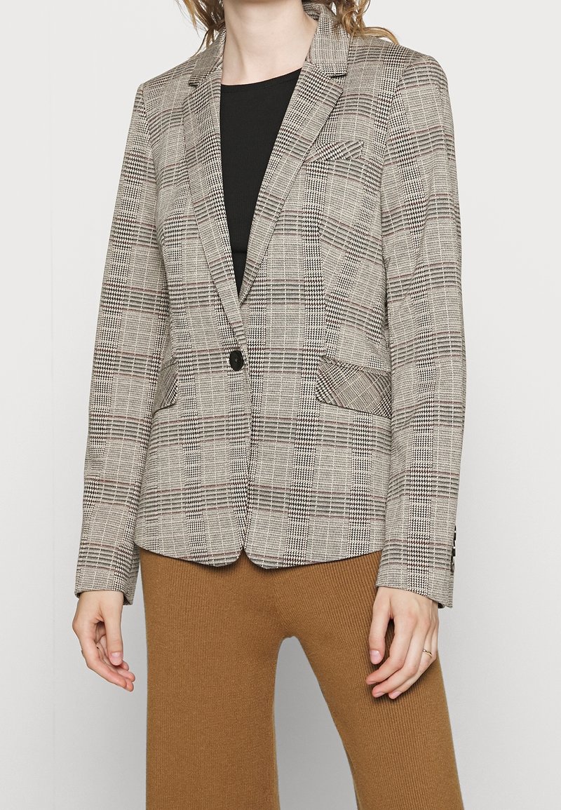 Personne portant un blazer à carreaux beige et marron avec un bouton noir unique et un pantalon côtelé couleur camel, sur un fond uni.