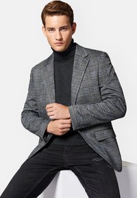 Szary marynarka w kratę z teksturowanej tkaniny z niebieskimi akcentami, noszona na czarnym golfie. Model siedzi, dłonie złożone przed sobą.