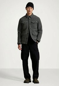 Άνδρας που φορά γκρι πουκαμίσα με κουμπιά, μαύρο παντελόνι cargo, μαύρη μπαντάνα, και ελιών παπούτσια, στέκεται μπροστά από ένα απλό φόντο.