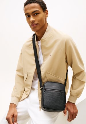 Junger Mann mit beiger Jacke und weißer Hose, der eine schwarze Tommy Hilfiger Umhängetasche mit strukturiertem Vorderteil trägt.