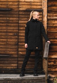 Black long coat made of fabric with a high collar, zip and snap closure, side pockets, and a brown logo patch. The person wears black pants and boots.