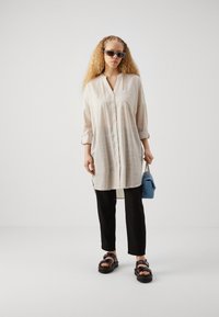 Chemise longue beige à rayures verticales, avec un devant à boutons et des manches retroussées, assortie à un pantalon noir et des sandales épaisses noires. Sac à main bleu inclus.