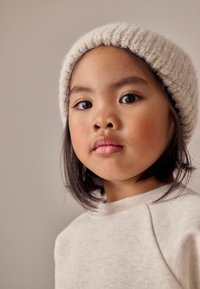 Beige ribbad stickad mössa på barn med axellångt svart hår. Ljusgrå sweatshirt med rund halsringning och mjuk textur.