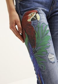 Blue denim jeans featuring a large, colorful parrot design with green leaves, distressed areas, and detailed stitching on the thigh.