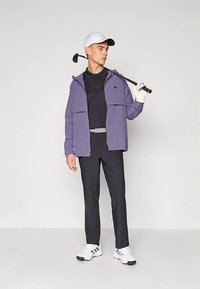 Golfa apģērbā ir gaiši violetas krāsas džempera jaka ar rāvējslēdzēju, melna polo krekls, melnas bikses, baltas kurpes un balta cepure, pie pleca pārnests golfa klubs.