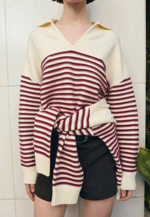 Sweter w paski z dzianiny w kolorze kremowym i bordowym, z dekoltem w serek, oversize'owymi rękawami oraz wiązanym detalem z przodu, zestawiony z ciemnymi szortami.