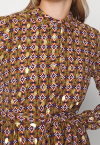 Geometriskt mönstrad klänning i röd, blå, guld och krämfärgad nyanser. Tyget har en mjuk textur med krage och insvängd midjedetalj.