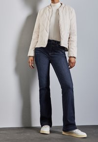 Chaqueta bomber de crema acolchada con puños acanalados, usada sobre una blusa clara, combinada con jeans de campana azul oscuro y zapatillas blancas.