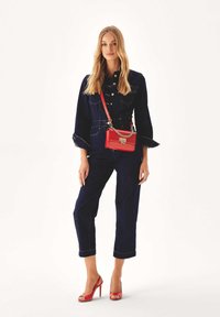 Denim jumpsuit dengan kain biru, tombol besar, dan lengan mengembang. Dipasangkan dengan tas selempang merah dan sepatu hak terbuka merah.