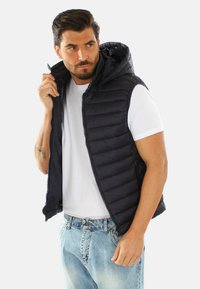 Gilet imbottito nero con cappuccio, caratterizzato da una chiusura con cerniera e un design trapuntato orizzontale. Indossato sopra una maglietta bianca e jeans blu.