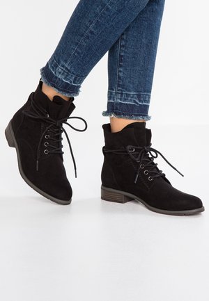 Czarne zamszowe botki na sznurowadła noszone z niebieskimi dżinsami z postrzępionymi nogawkami na białym tle.