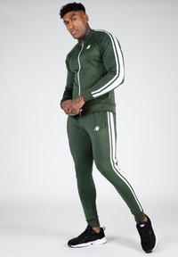 Grünes sportliches Trainingsanzug mit weißen Seitenstreifen und Logo. Schlanker Schnitt, lange Ärmel, Reißverschluss vorne, Material wirkt glatt und dehnbar. Schwarze Turnschuhe.