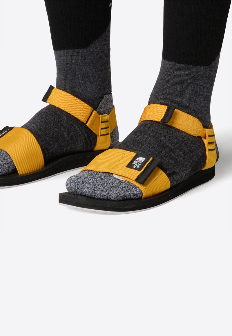 Gula sandaler med justerbara remmar, som bärs över mörkgrå stickade strumpor. Sandalerna har en texturerad sula och en svart yttersula.