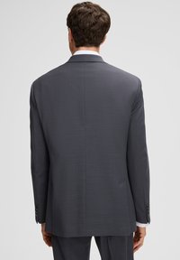 Mężczyzna ubrany w ciemnoszare teksturowane marynarkę i dopasowane spodnie, stojący tyłem na tle jednolitego jasnoszarego tła.