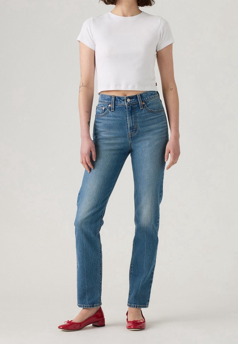 Femme portant un jean bleu taille haute, un t-shirt blanc court et des chaussures rouges à talons bas, debout devant un fond uni.