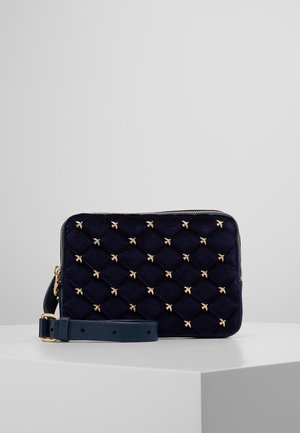 Pochette bleu marine en velours matelassé avec ornements en forme d'abeille dorée et bracelet en cuir amovible, présentée sur un socle blanc.