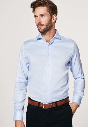 Light blue, long-sleeve shirt with a pointed collar and button closure. Smooth fabric with a slight sheen. Worn with a brown belt and dark pants.