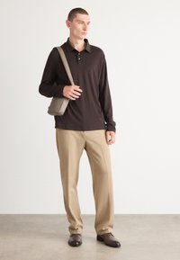 Hosszú ujjú sötétbarna polo ing, bézs nadrág és barna cipő. A modell egy világosszürke crossbody táskát tart, és egy minimalista háttér előtt áll.