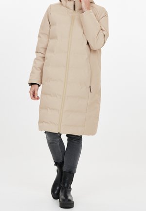 Personne portant un manteau long matelassé beige avec capuche, un jean skinny gris et des bottines noires, debout devant un fond blanc.