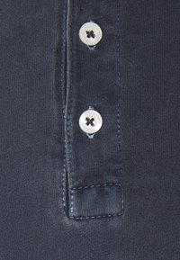 Mörkblå polo-tröja med fint tyg, prydd med två vita knappar och en liten broderad lapp. Ren, minimalistisk design.