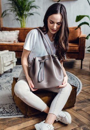 Bucket bag in smooth taupe leather with gray drawstring closure. Set against a wooden floor and a plush brown sofa, featuring a logo accent.