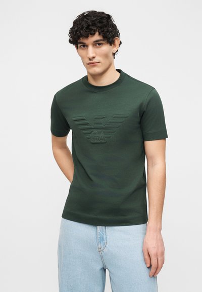 Νεαρός με κατσαρά μαλλιά φοράει σκούρο πράσινο κοντομάνικο μπλουζάκι με ανάγλυφο λογότυπο και ανοιχτό μπλε τζιν, στέκεται μπροστά σε απλό φόντο.