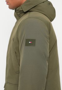 Férfi olíva zöld párnázott dzsekit visel, kapucnival és Tommy Hilfiger logóval a vállán, oldalnézet.