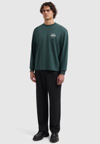 Man met krullend haar die een donkergroen langemouwen shirt met een klein wit logo en zwarte broek draagt, staand tegen een effen lichte achtergrond.