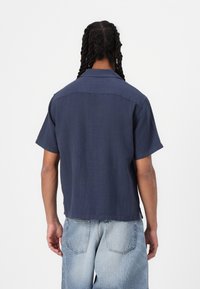 Camisa de manga corta en tela ligera color marino, con dobladillo recto, cuello abotonado y un corte relajado, combinada con unos jeans de mezclilla claros.