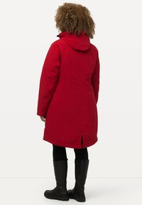 Red padded coat with a hood, long sleeves, side slits, and a smooth texture. Worn over black pants and tall black boots.