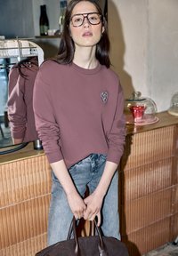 Jeune femme aux longs cheveux et lunettes, portant un sweat-shirt mauve avec un patch en forme de cœur, un jean en denim clair, tenant un sac en cuir marron foncé à l'intérieur.