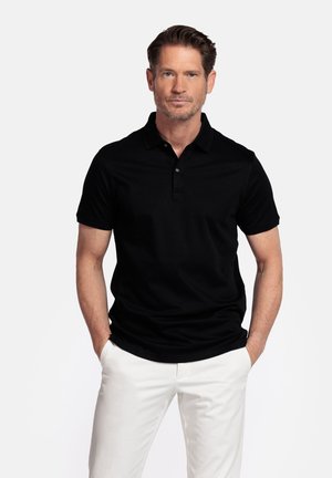 Man staat met handen in zakken, draagt een zwarte korte mouw polo en witte broek tegen een effen witte achtergrond.