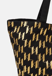 Bolso de mano negro con un patrón geométrico dorado en toda la superficie y texto de la marca. Fabricado con un tejido resistente y con dos asas.