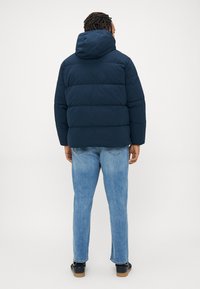 Navy puffer jacket with a hood, featuring quilted sections; paired with light blue jeans and dark shoes, viewed from the back.