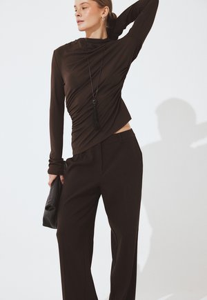 Žena pózuje v priliehavom hnedom dlhom rukáve s riasením, vo voľných hnedých nohaviciach, nesie čiernu listovú kabelku a dlhý čierny náhrdelník s strapcami.