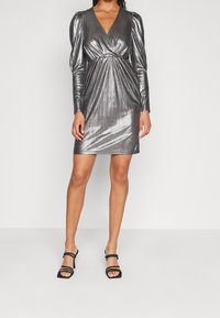Metallisk silvrig klänning med långa puffärmar, v-ringad halsringning och samlad midja. Buren med svarta remsandaler som framhäver en slank silhuett.