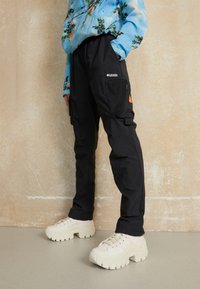 Person med svarta Columbia cargo-byxor med nätficka och kraftiga vita sneakers, som står mot en strukturerad beige vägg.