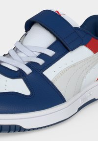 Λευκό, μπλε και κόκκινο αθλητικό παπούτσι Puma με λευκά κορδόνια, μπλε ιμάντα, υφασμένο πάτο και λογότυπο Puma στο πλάι και στη γλώσσα του παπουτσιού.