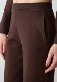 Pantaloni lounge marroni realizzati in un tessuto texturizzato, con una vita ampia e tasche laterali. I pantaloni hanno un aspetto morbido e accogliente.
