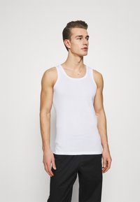 Hvid bomulds tanktop med rund halsudskæring og brede skulderstropper, båret med sorte bukser, der har en glat overflade.